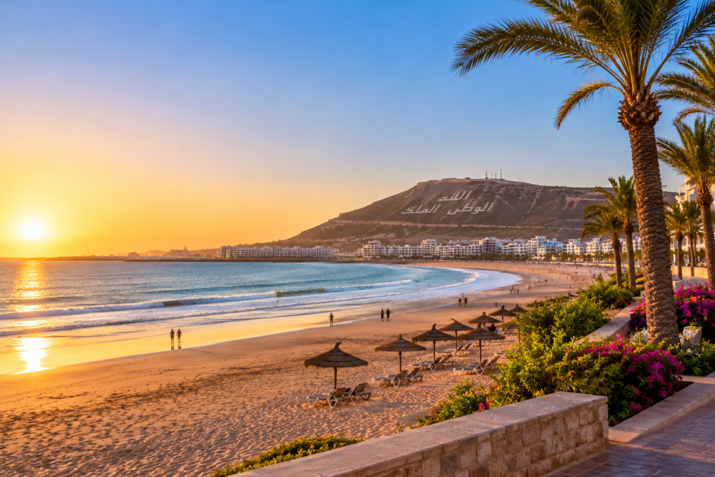 Plage d’Agadir au coucher du soleil, avec palmiers, parasols et la colline de la kasbah